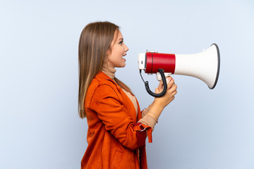 Teenager girl with coat over isolated blue background shouting through a megaphone