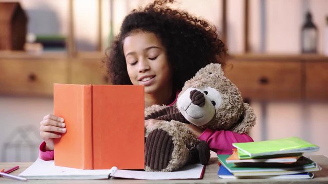 smiling african american child reading book to teddy bear
