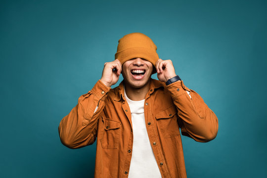I can't see you! Photo of attractive mixed race man in yellow outfit, wearing hat and glasses smiling in good mood isolated over blue background, pull hat over his head and laugh out loud