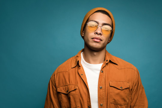 Photo Of Good-looking Confident Mixed Race Man In Yellow Outfit, Wearing Hat And Glasses Looking Straight At Camera, Isolated Over Blue Background