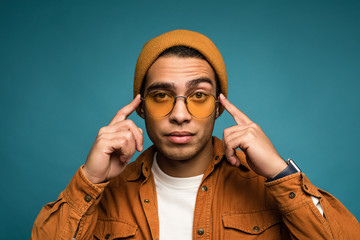 Photo of attractive concerned mixed race man in yellow outfit, wearing hat and glasses. slightly touching his temples, isolated over blue background