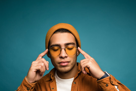 Photo of handsome composed mixed race hipster man in yellow outfit, wearing hat and glasses, touching his forehead with hands, isolated over blue background