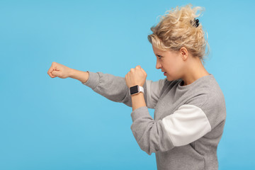 Fototapeta na wymiar Ready to fight! Side view of feisty woman with short curly hair in sweatshirt punching to side with aggressive angry expression, knockout with fist. indoor studio shot isolated on blue background