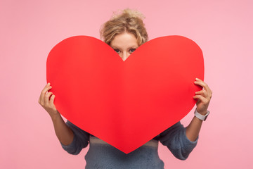 Portrait of young woman with curly hair hiding face behind big red heart and looking at camera with...