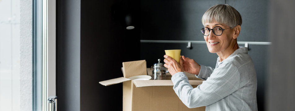 Panoramic Shot Of Smiling Mature Woman Unpacking Box In New House