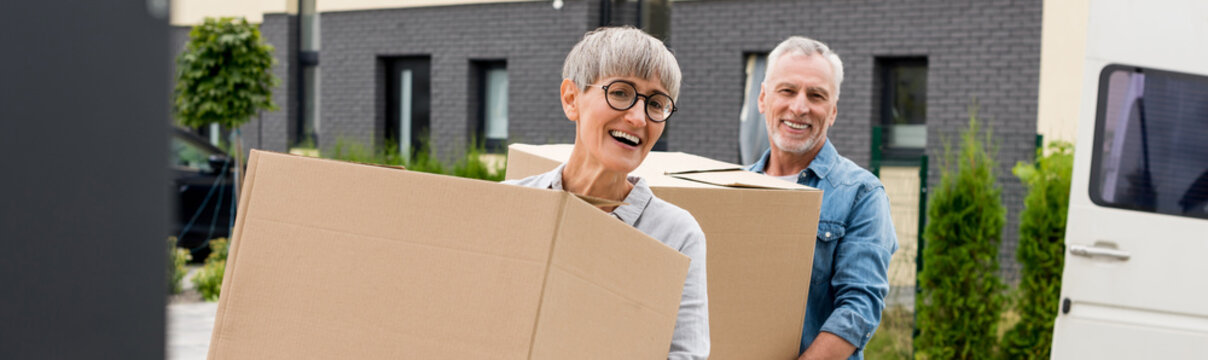 Panoramic Shot Of Mature Man And Smiling Woman Bringing Boxes To New House