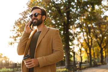 Image of handsome adult man talking on smartphone while walking in park