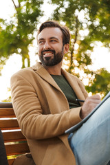 Fototapeta premium Image of handsome man woking and holding clipboard on bench in park