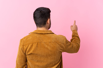 Young handsome man with beard wearing a corduroy jacket over pink background pointing back with the...