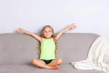 Joyful pretty little girl with beautiful long hair sitting on cozy sofa with raised hands and smiling looking at camera, child relaxing on couch enjoying childhood, feeling carefree and happy. indoor