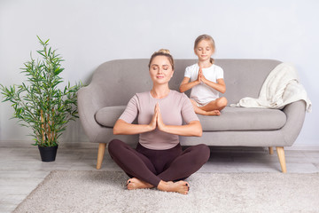 Fototapeta na wymiar Lotus posture. Young mother and little daughter with closed eyes and prayer gesture practicing yoga together at home, doing exercise breath technique, meditating in room, mindfulness and harmony