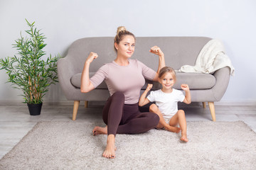 Attractive fit mom and her charming daughter showing biceps, smiling at camera, sitting together on...