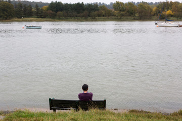 Fototapeta na wymiar A lone man sitting isolated on a bench of the bank of a river