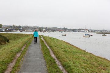 A lone middle aged woman taking a walk along a river at high tide on a dull and rainy day