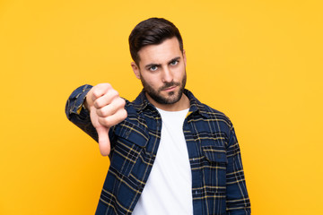 Young handsome man with beard over isolated yellow background showing thumb down with negative...