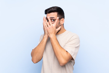 Young handsome man with beard over isolated blue background covering eyes and looking through fingers