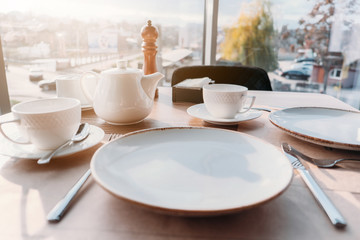 Table setting for breakfast, lunch in sun light. Dinner plate setting with empty plates, cups, teapot near window in cafe, restaurant