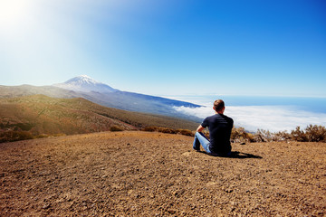 Naklejka na ściany i meble A young male traveler sits on the sand and looks at the top of the Teide volcano. Teide National Park, Canary, Spain