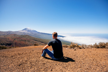 A young male traveler sits on the sand and looks at the top of the Teide volcano. Teide National Park, Canary, Spain