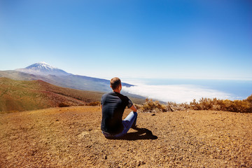 A young male traveler sits on the sand and looks at the top of the Teide volcano. Teide National Park, Canary, Spain