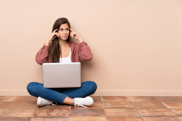 Teenager student girl sitting on the floor with a laptop having doubts and thinking