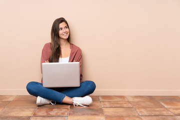 Teenager student girl sitting on the floor with a laptop looking up while smiling