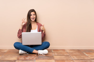 Teenager student girl sitting on the floor with a laptop showing ok sign and thumb up gesture