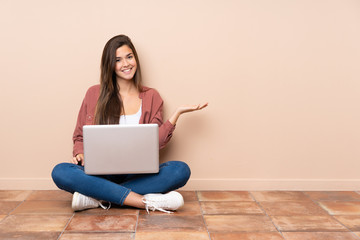 Teenager student girl sitting on the floor with a laptop holding copyspace imaginary on the palm to insert an ad