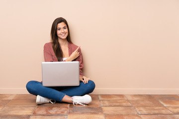 Teenager student girl sitting on the floor with a laptop pointing to the side to present a product