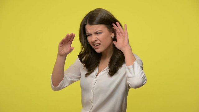 What, i can't hear you! Irritated woman with brunette hair in blouse holding hand near ear and pretending not to listen, showing bla bla gesture, uninterested in talk. studio shot, yellow background