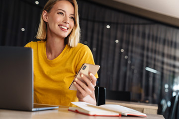Photo of smiling blonde woman using laptop and smartphone while sitting