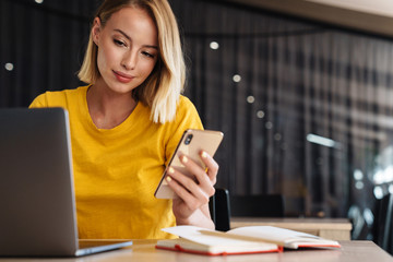 Photo of young blonde woman using laptop and smartphone while sitting