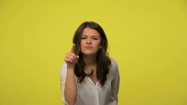 Frustrated angry brunette woman in blouse showing lie gesture, pointing nose and at camera, meaning you are liar, dishonest person. falsehood fake information. indoor studio shot, yellow background