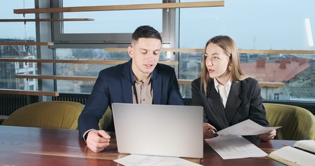 Man and a woman discussing work in the brightly lit modern office. Concerned male and female working with laptop and charts papers. Business people discuss new startup project concept. Moving camera
