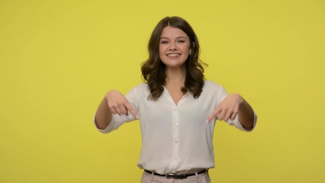 Friendly cheerful brunette woman pointing down and smiling at camera, meaning subscribe or follow information below, showing place for promotion text, advertising area. studio shot, yellow background