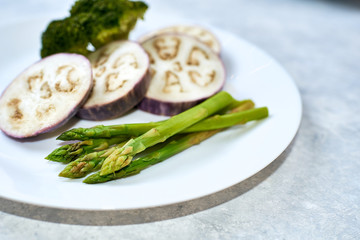 Healthy eating concept. Steamed vegetables in a white plate on a blue table. Eggplant, broccoli, asparagus.