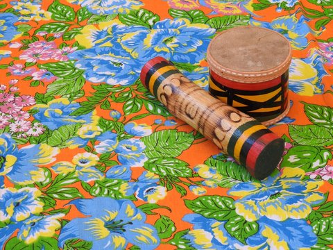 Two colorful handmade Brazilian percussion instruments: small drum and "ganz&aacute;", a type of rattle, on a colorful &ldquo;chitao&rdquo; fabric with large floral prints. They are widely used in axe music.