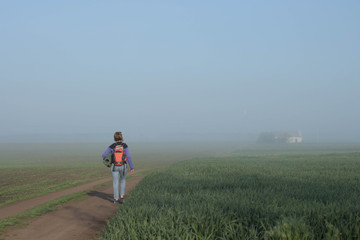 Woman traveler with backpack walks in beautifyl atmospheric misty morning  a house among fields in the background