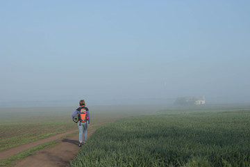 Woman traveler with backpack returns home in beautifyl atmospheric misty morning  a house among fields in the background