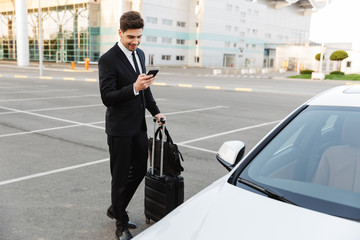 Image of young caucasian businesslike man standing with travel luggage and cellphone by car outdoors