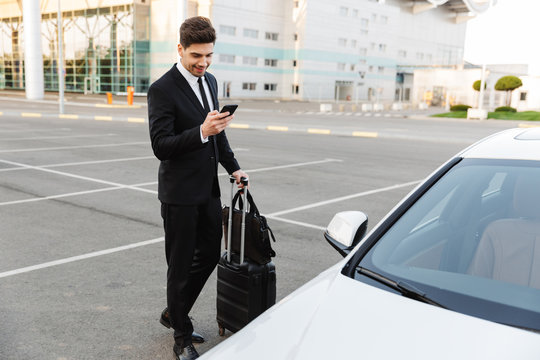 Image of young caucasian businesslike man standing with travel luggage and cellphone by car outdoors