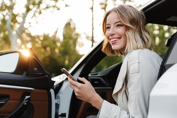 Image of beautiful businesslike woman sitting in car and using cellphone - obrazy, fototapety, plakaty