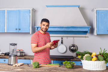 Positive man standing in modern kitchen holding tablet pc and looking at camera with smile, searching food recipe on internet, reading calories, basket of fresh green vegetables on table, healthy diet