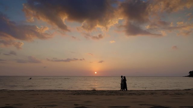 Silhouettes of adult couple are walking in water edge on sunset tropical beach with dramatic colorful sky and jet ski in sea