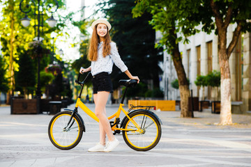Obraz na płótnie Canvas Girl teenager in a hat with a yellow bike walks through the summer city.