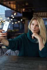 Blonde Caucasian woman in green pullover making selfie on her smartphone while waiting for her friend nothing to do more but photo herself using device as mirror