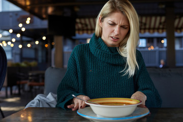 Blonde Caucasian woman in green pullover demonstrates disgust twisting face with negative reaction while trying to eat some smelly soup from a plate on a table