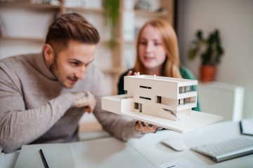Architects with model of house sitting at the desk indoors in office, working.