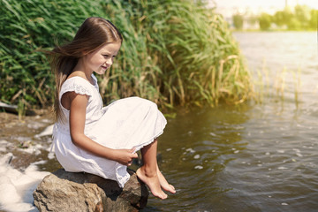 cute little girl in a white dress sits on a lake on a stone