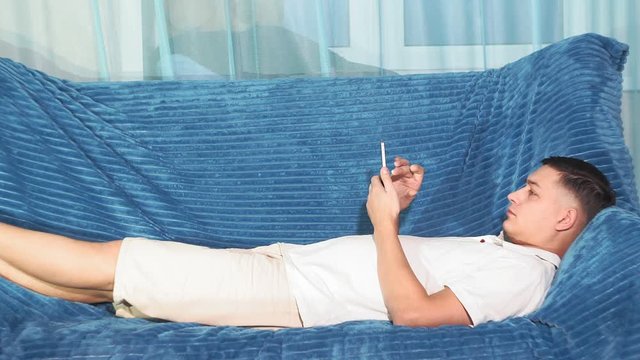 young man in a white t-shirt is lying on the sofa with a phone